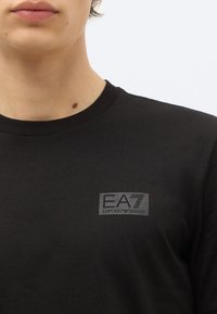 Osoba nosi čierne tričko s okrúhlym výstrihom a malým sivým logom EA7 Emporio Armani na ľavej strane hrudníka.