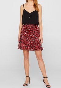 Zwarte mouwloze top met knopen, gecombineerd met een rode rok met ruffles en een zwart luipaardprint ontwerp. Dunne zwarte enkelbandjes op de schoenen.