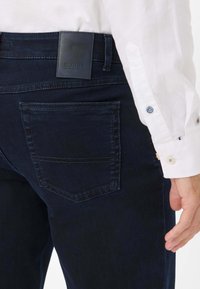 Donkerblauwe spijkerbroek van denim met een achterzak en een leren label met de tekst "EUREX." Het materiaal is soepel met zichtbare stikseldetails.