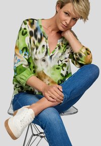 Frau mit kurzen blonden Haaren, die eine grüne gemusterte Bluse, blaue Jeans und weiße Sneakers trägt, sitzt mit einem Bein über das andere geschlagen auf einem Drahtstuhl.