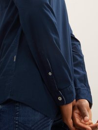 Marineblaues Hemd mit Knöpfen, Kragen, langen Ärmeln und seitlichen Schlitzen. Betont eine glatte Stoffstruktur und Knopfdetails an den Manschetten.