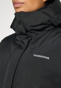 Close-up of a person wearing a black Didriksons jacket with a high collar zipped up near the chin.