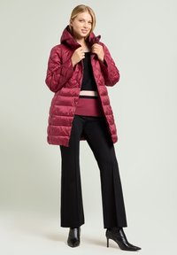 Weinroter Stepp-Puffer-Mantel mit Kapuze, gesteppter Optik und figurbetonter Silhouette. Getragen über einem schwarzen Oberteil und weinroter Hose, kombiniert mit schwarzen High Heels.