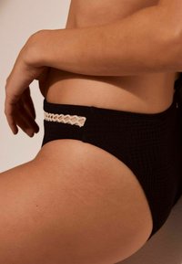 Melna bikini apakša ar faktūras audumu, ar bežīgu ģeometrisku akcentu gar jostasvietu, izceļot piegulošu dizainu.