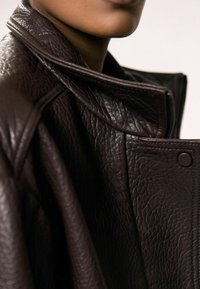 Giubbotto in pelle marrone con una finitura testurizzata, caratterizzato da un colletto ripiegato e una chiusura con zip. La vista ravvicinata enfatizza la qualità del materiale.