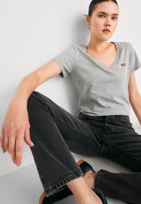 Szürke V-nyakú póló, egy kis piros Levi's logóval, fekete farmernadrággal párosítva. A modell egy fehér háttér előtt ül, kék cipőt visel.