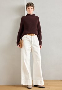 Maglione a collo alto a coste marroni, pantaloni a gamba larga crema, cintura color cammello, in mano una pochette color cammello e stivaletti beige con tacco.