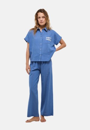 Femme portant une chemise bleue ample boutonnée avec une broderie de poissons et un pantalon à jambes larges assorti, debout pieds nus, une main sur la hanche.