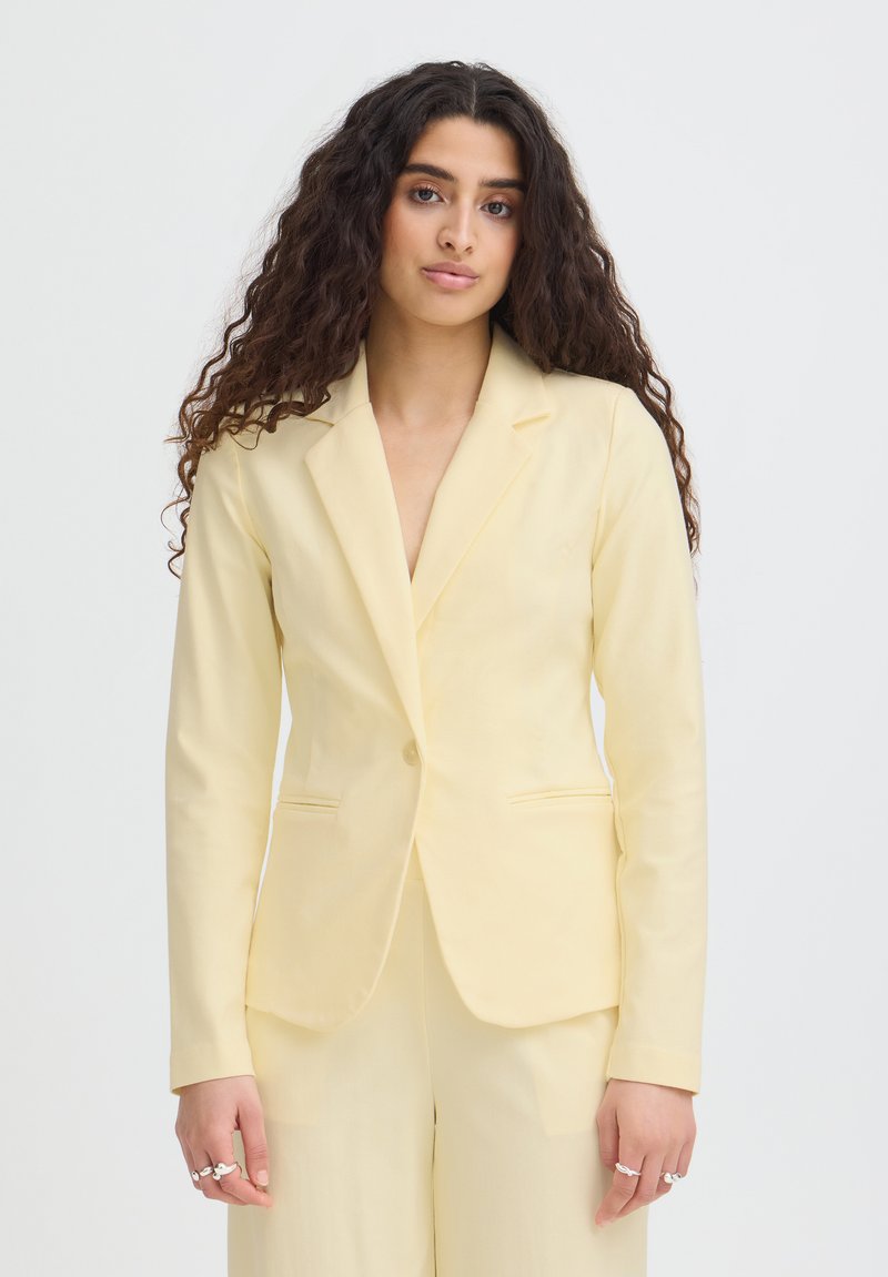 Femme aux longs cheveux bouclés portant un blazer jaune pâle ajusté et un pantalon assorti, debout sur un fond blanc.