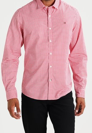 Mężczyzna ubrany w czerwoną i białą koszulę w kratę typu gingham z długim rękawem oraz czarne spodnie, stojący na jednolitym tle.