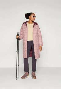 Lys rosa quilted lang frakk med mørk lilla fôr, beige topp, mørke bukser og brune sko. Personen holder en svart gangstokk.