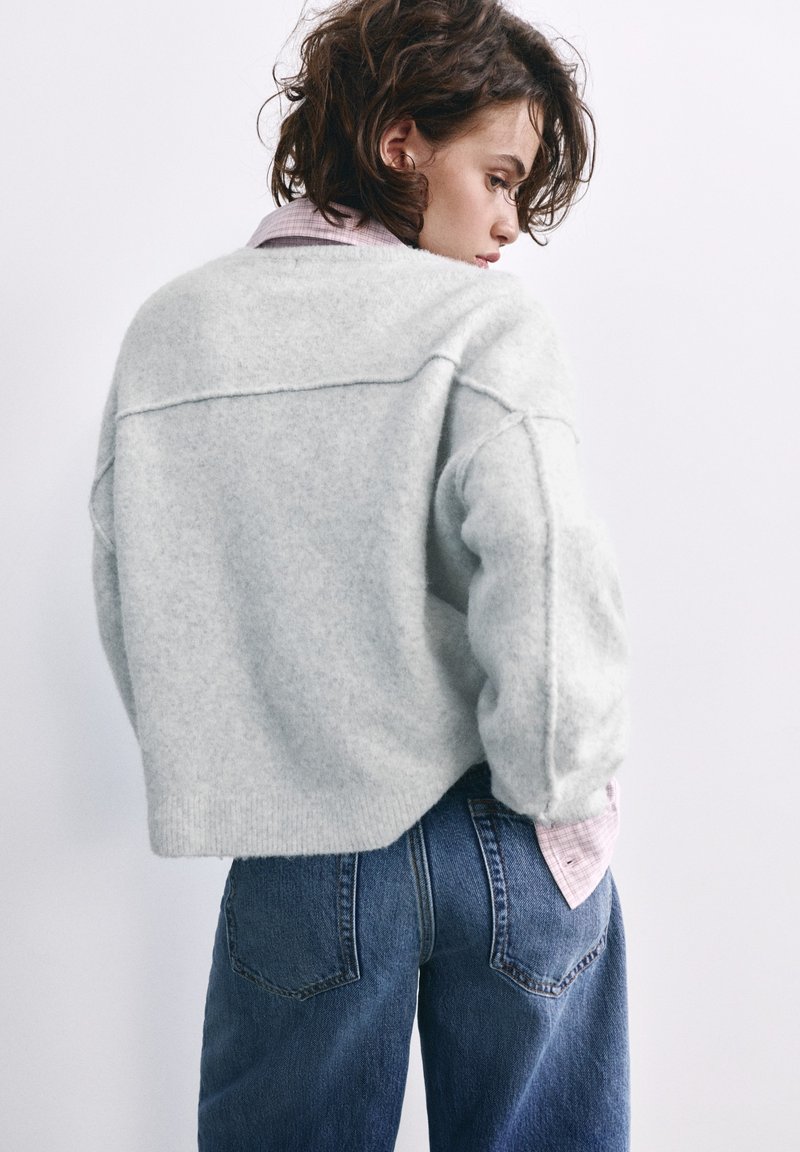 Femme aux cheveux courts et bouclés portant un pull pelucheux gris clair sur une chemise rose et un jean bleu, de dos avec la tête tournée sur le côté.
