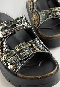 Sandalias negras con tiras adornadas que presentan cuentas plateadas, doradas y negras, lentejuelas metálicas y cierres de doble hebilla. Suela plana de corcho.