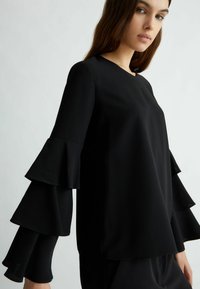 Maglietta nera a maniche lunghe con dettagli a volants arricciati sulle maniche, tessuto morbido e scollatura rotonda. Design semplice e classico.