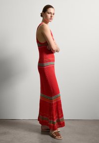 Rotes gehäkeltes Maxikleid mit horizontalen Streifen in Grün und Pink, das eine figurbetonte Silhouette und einen offenen Rücken hat. Kombiniert mit braunen Sandalen mit Riemchen.