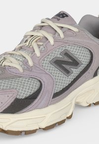 Κοντινό πλάνο ενός γκρι και λεβαντίν New Balance αθλητικού παπουτσιού με λευκά κορδόνια και υφασμάτινη πλευρική επιφάνεια με το λογότυπο της μάρκας.