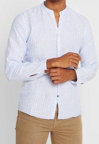 Chemise bleu clair à manches longues avec un col mao, des rayures verticales et une fermeture avant par boutons. Tissu doux et texturé avec des boutons contrastants.