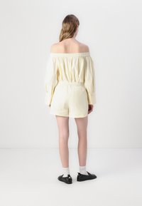 Off-shoulder, lysegul romper med elastisk midje og korte ermer, laget av glatt stoff og med avslappet passform, parret med svarte sko.