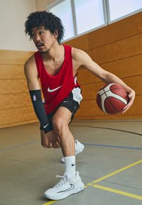 Punane Nike triiksärk, mustad lühikesed püksid, valged tossud ja must käepide, millel on NBA logo. Mängija dribblib korvpalli spordisaali põrandal.