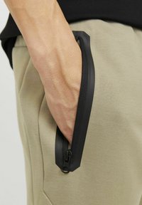 Beige pants with a black zippered pocket, featuring a textured fabric and a distinctive black zipper with a rounded pull tab.
