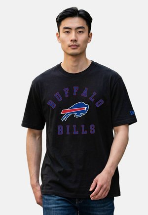 Mężczyzna stojący twarzą do przodu, ubrany w czarną koszulkę Buffalo Bills i niebieskie dżinsy na jednolitym jasnym tle.
