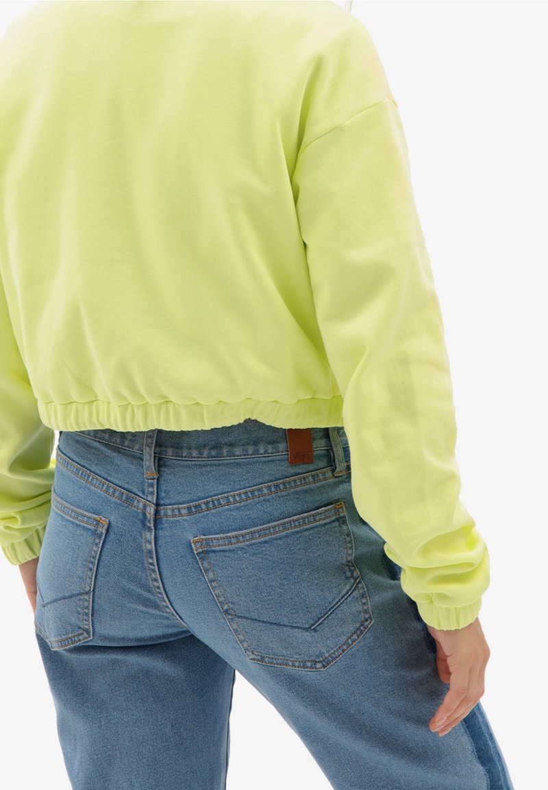 Sudadera amarilla neón ajustada con dobladillo elástico y mangas largas, combinada con jeans de mezclilla azul de talle alto con bolsillos traseros.