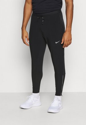 Mężczyzna w czarnych spodniach dresowych Nike ze sznurkiem i białych butach sportowych Nike, stojący na jednolitym białym tle.