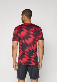 Rot-schwarz gestreiftes Sportshirt mit kurzen Ärmeln, aus atmungsaktivem Stoff, getragen mit grauen Shorts.