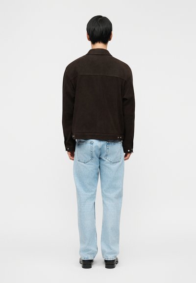 Άνδρας με καφέ σκούρο μπουφάν, ανοιχτό μπλε τζιν και μαύρα παπούτσια, που κοιτάζει προς τα πίσω, μπροστά σε λευκό απλό φόντο.