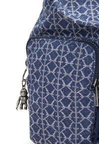 Denim backpack with a geometric white pattern, featuring a zippered pocket and a silver figurine charm attached to the zipper.