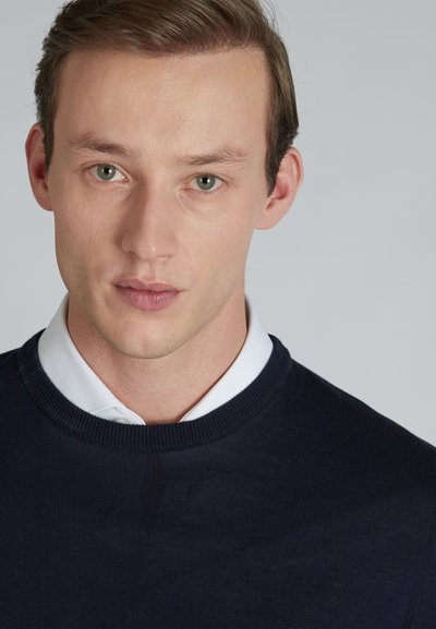 Navy sweater with a round neckline, textured knit, worn over a white collared shirt. Smooth fabric with subtle stitching details.