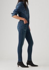 Mörka skinny jeans i denim med hög midja, kombinerade med en denimskjorta. Modellen bär svarta ankelstövlar, med händerna i fickorna.
