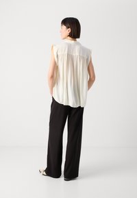 Krämlik ärmlös blus med draperad ryggdetalj, kombinerad med svarta vida byxor och platta skor, visad bakifrån mot en ljus bakgrund.