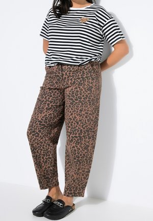 Femme debout, les mains derrière le dos, portant un haut rayé noir et blanc avec un patch léopard, un pantalon imprimé léopard, et des chaussures noires sans lacets.