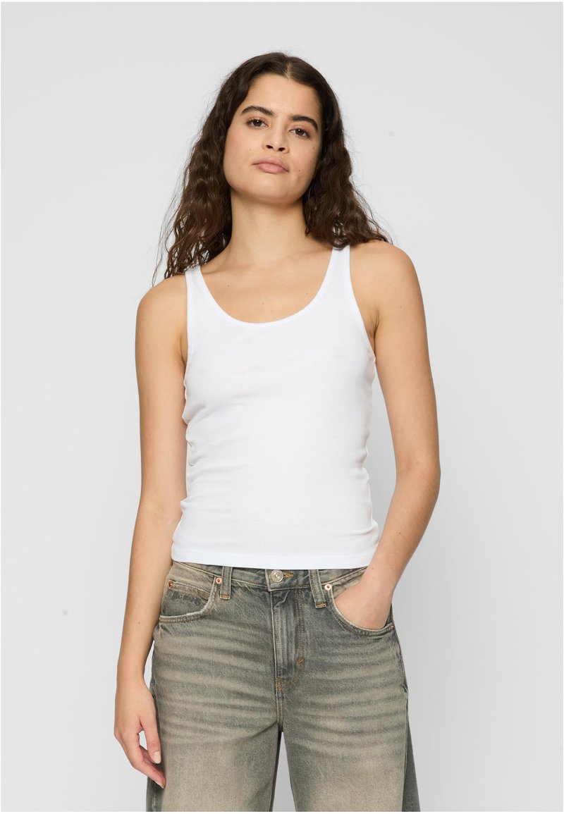 Canotta di cotone bianca con scollatura a girocollo, caratterizzata da spalline sottili e un design attillato, indossata con jeans larghi e sbiaditi.