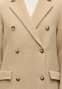 Manteau en laine beige croisé avec six boutons et larges revers, porté sur un haut en maille blanc.