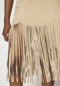 Bézs suédból készült francia szoknya, amelynek ívelt szegélyéről hosszú, vékony fröccsök lógnak. Díszvarrás található a felső részén.