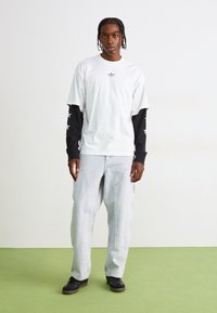 Tricou alb suprapus peste o bluză neagră cu mânecă lungă, cu accente în formă de stea, asortat cu blugi gri deschis, cu croială lejeră și adidași negri.