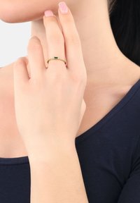 Anello d'oro con una pietra verde a taglio smeraldo, accentuato da piccole pietre trasparenti lungo la fascia; indossato su una mano che tocca un collo.
