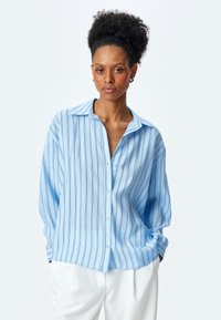 Camisa de botones azul claro con rayas verticales, corte relajado y textura suave; cuenta con cuello y mangas largas, combinada con pantalones blancos.