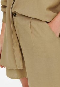 Detailní záběr osoby nosící béžové lněné šortky s knoflíkem a rukou v boční kapse, doplněné o odpovídající košili.