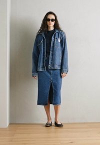 Conjunto de chaqueta y falda de mezclilla, azul medio, con un corte holgado y una abertura en la parte delantera de la falda. Top de punto texturizado. Gafas de sol negras y zapatos planos.