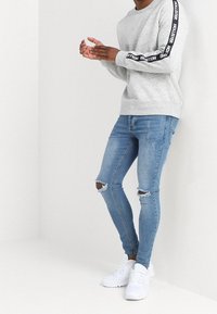 Person i ljusgrå sweatshirt med svarta "Hollister"-ränder på ärmarna, trasiga ljusblå skinny jeans och vita sneakers, som står mot en vägg.