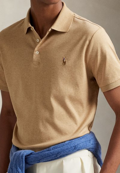 Prim-plan cu o persoană care poartă un tricou polo bej cu un mic logo brodat și un pulover albastru legat în jurul taliei.