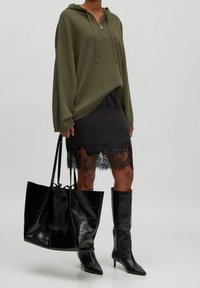 Sweat à capuche surdimensionné vert olive avec fermeture éclair, superposé sur une jupe noire à ourlet en dentelle. Sac cabas en cuir verni noir et bottes noires montantes jusqu'aux genoux.