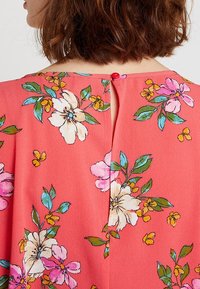 Korallefarbenes Blusenshirt mit floralen Mustern in Pink, Weiß und Gelb. Knopfschluss hinten mit einem kleinen Schlüssellochdetail. Weiche Stofftextur.