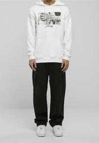 Sudadera blanca con capucha y un estampado gráfico que muestra manos y tatuajes. Cuenta con un bolsillo frontal, capucha con cordón y puños acanalados. Combinada con pantalones negros.