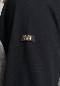 Schwarzes Langarmkleidungsstück mit einer weichen Stoffoberfläche und einem auffälligen Etikett mit goldfarbener Stickerei, das sich auf dem linken Ärmel befindet.