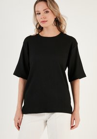 Black ribbed t-shirt with short sleeves and a round neckline, made from textured fabric. Worn with light-colored pants.
