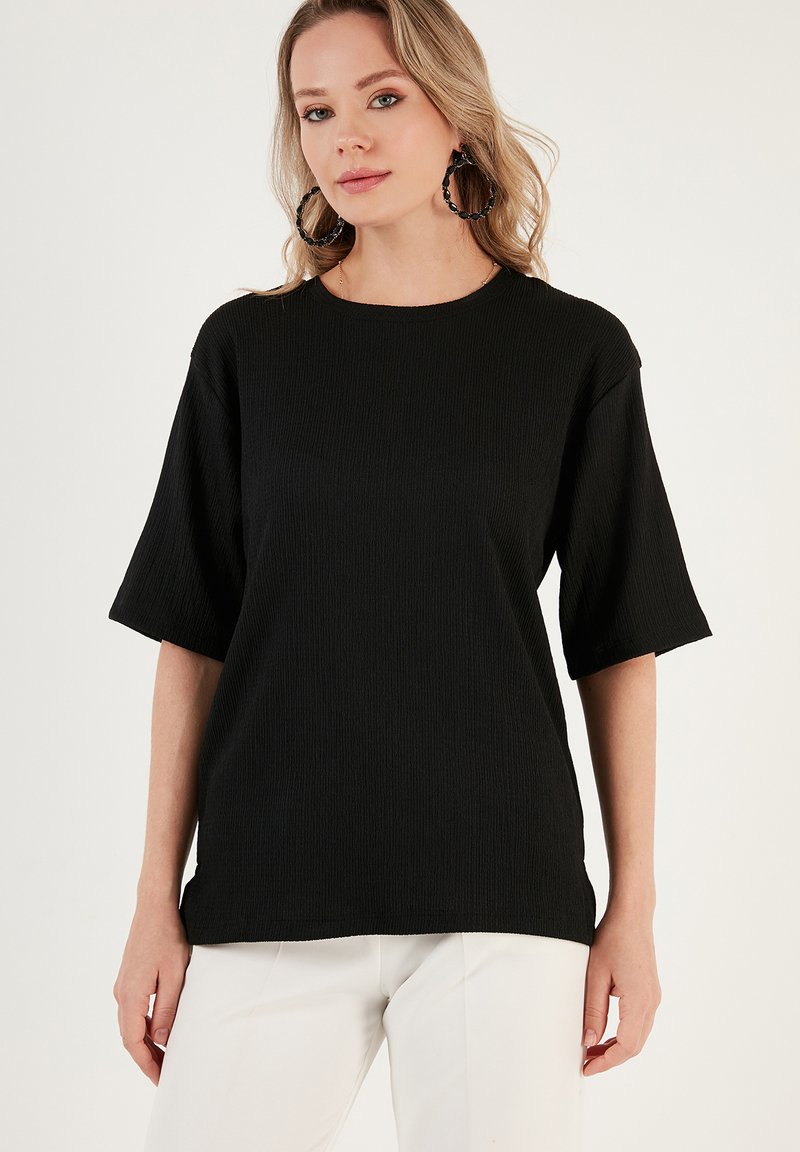 Black ribbed t-shirt with short sleeves and a round neckline, made from textured fabric. Worn with light-colored pants.